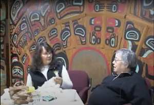 Two elders in conversation in front of carved formline screen