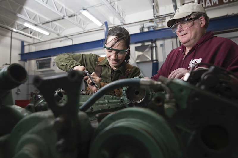 Instructor oversees a student working on an engine block